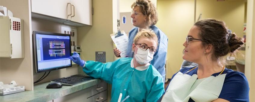 Two dental hygiene students and an instructor in their uniforms going over test results on a computer screen.